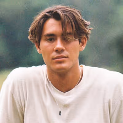 Young man with wavy brown hair wearing a white t-shirt and a pendant necklace, posing outdoors with a blurred green background.